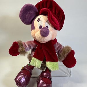 May include: A plush Minnie Mouse toy wearing a deep red velvet hat, a matching red scarf with light green ends, a mauve velvet jacket with brown faux fur cuffs, and shiny dark purple shoes. A tag reads Disneyland Resort Paris.