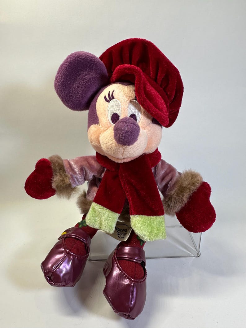 May include: A plush Minnie Mouse toy wearing a deep red velvet hat, a matching red scarf with light green ends, a mauve velvet jacket with brown faux fur cuffs, and shiny dark purple shoes. A tag reads Disneyland Resort Paris.