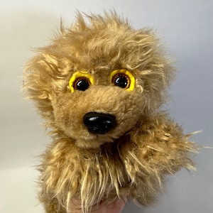 Puede incluir: Un peluche con pelaje largo y desordenado de color marrón claro. La criatura presenta grandes ojos de color marrón oscuro rodeados por anillos de tela de color amarillo brillante y una nariz negra lisa de forma ovalada.