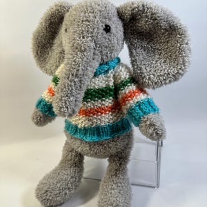 Puede incluir: Un juguete de peluche de elefante gris con textura difusa, que presenta orejas grandes y una trompa. El elefante lleva un suéter tejido con rayas horizontales en blanco, naranja y verde, terminado con un cuello y puños acanalados turquesa.