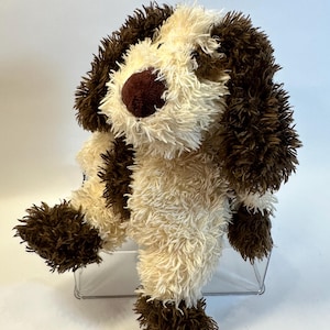 May include: A plush dog toy featuring shaggy cream and dark brown fur. The stuffed animal has long, floppy dark brown ears, a dark brown nose, and a seated posture. The texture of the fur is long and slightly tousled throughout the body.
