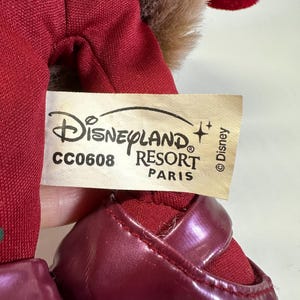 May include: A plush toy featuring a red fabric body and dark red shiny shoes. A cream-coloured tag is attached, displaying the text Disneyland Resort Paris, the code CC0608, and the Disney copyright symbol. Brown faux fur trim is visible at the top.
