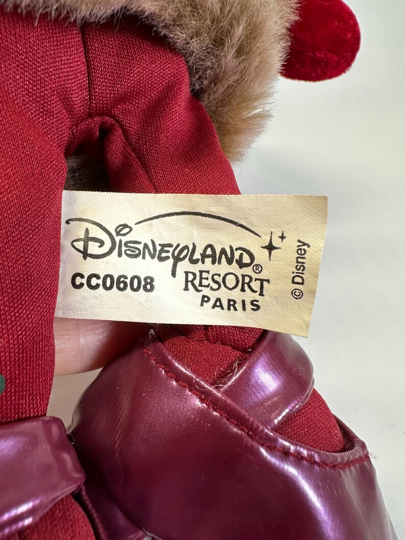 May include: A plush toy featuring a red fabric body and dark red shiny shoes. A cream-coloured tag is attached, displaying the text Disneyland Resort Paris, the code CC0608, and the Disney copyright symbol. Brown faux fur trim is visible at the top.