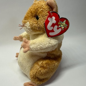 Peut inclure: Une peluche en forme de hamster avec une fourrure dorée et crème, dotée de petites pattes roses et d'une étiquette rouge en forme de cœur attachée à l'oreille, affichant les lettres blanches ty et une étoile holographique avec le mot BEANIES.