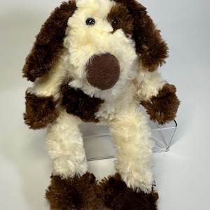 May include: A plush dog toy featuring soft, cream-coloured fur with dark brown patches on the ears, body, and feet. The stuffed animal has a prominent, rounded dark brown nose, two small black eyes, and long, floppy ears that hang down the sides.