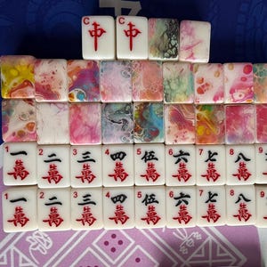 May include: A collection of white Mahjong tiles with red Chinese characters and colorful abstract designs. The tiles are arranged in rows, displaying various suits and numbers. The top row features the "C" and "中" tiles.