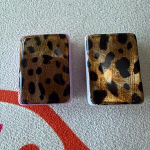 May include: Two rectangular tiles featuring a glossy leopard print pattern in shades of gold, brown, and black. The tile on the left has a light purple border, while the tile on the right has a white border. Both tiles have a smooth, reflective finish.