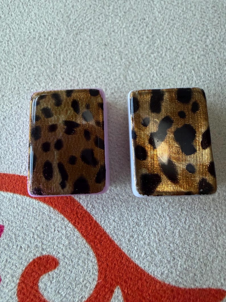 May include: Two rectangular tiles featuring a glossy leopard print pattern in shades of gold, brown, and black. The tile on the left has a light purple border, while the tile on the right has a white border. Both tiles have a smooth, reflective finish.