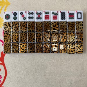 May include: A set of thirty Mahjong tiles arranged in three rows of ten. The top row features white tiles with colorful circular patterns and numbers one through nine, plus a blank tile. The remaining twenty tiles have a brown and black leopard print.