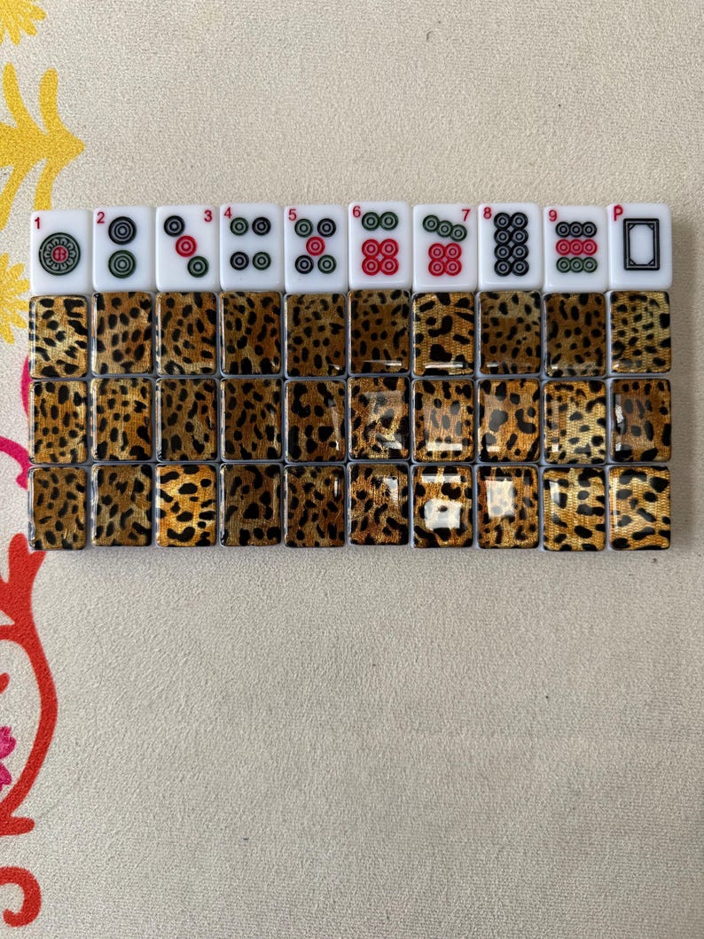 May include: A set of thirty Mahjong tiles arranged in three rows of ten. The top row features white tiles with colorful circular patterns and numbers one through nine, plus a blank tile. The remaining twenty tiles have a brown and black leopard print.