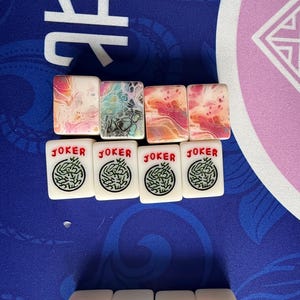 May include: A collection of Mahjong tiles. The top row features tiles with colorful abstract designs in pink, orange, and blue. The bottom row displays tiles with the word "JOKER" in red and a green circular design.