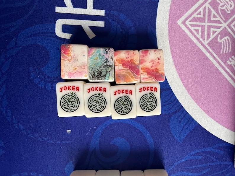 May include: A collection of Mahjong tiles. The top row features tiles with colorful abstract designs in pink, orange, and blue. The bottom row displays tiles with the word "JOKER" in red and a green circular design.