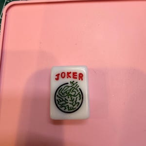 May include: A white rectangular mahjong tile featuring the word JOKER in red lettering above a circular black and green graphic design. The tile is positioned on a pink surface with a portion of a green measuring mat visible in the corner.