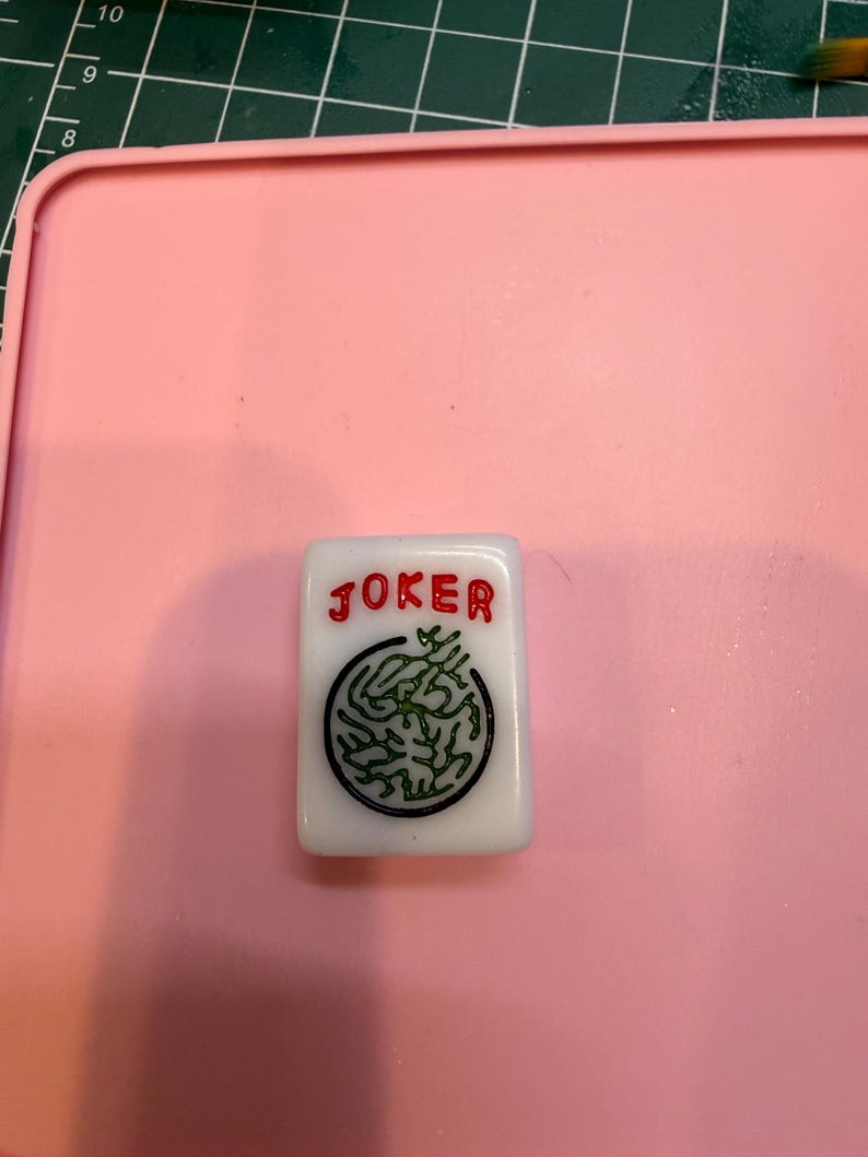 May include: A white rectangular mahjong tile featuring the word JOKER in red lettering above a circular black and green graphic design. The tile is positioned on a pink surface with a portion of a green measuring mat visible in the corner.
