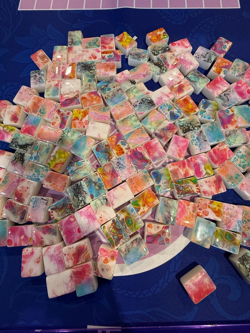 May include: A collection of small, rectangular, resin-coated tiles with colorful, abstract designs. The tiles feature a variety of colors, including pink, blue, orange, and white, with swirling patterns. The tiles are arranged on a blue surface.