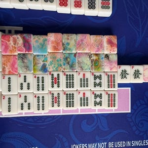 May include: A collection of Mahjong tiles arranged on a blue surface. The tiles feature a mix of white and colorful designs, including abstract patterns and traditional symbols. Some tiles display numbers and characters.