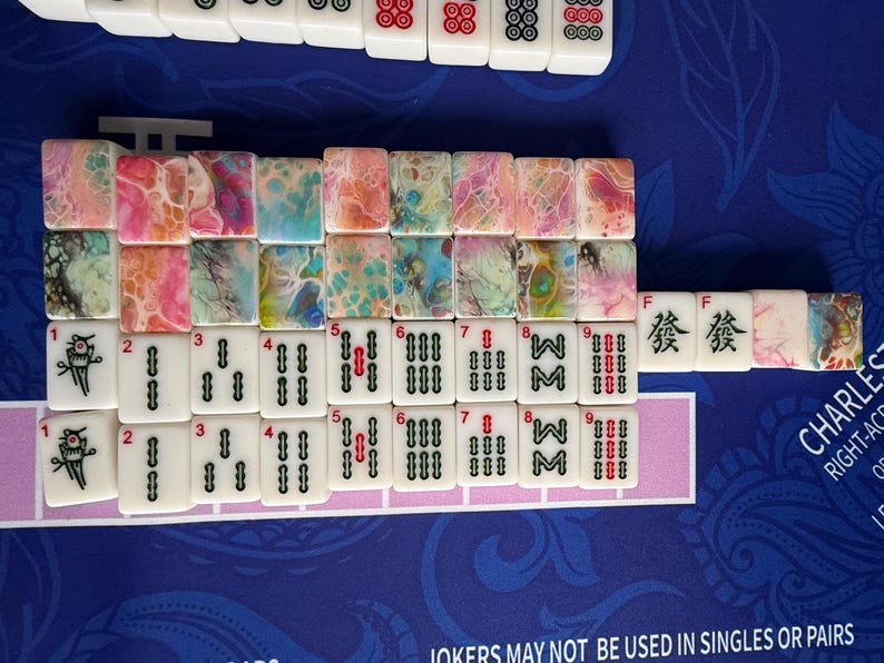 May include: A collection of Mahjong tiles arranged on a blue surface. The tiles feature a mix of white and colorful designs, including abstract patterns and traditional symbols. Some tiles display numbers and characters.