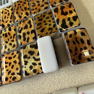 May include: A collection of rectangular mahjong tiles featuring a glossy leopard print pattern in shades of gold, brown, and black. Some tiles are solid white with black and red symbols, including numbers and letters, arranged in a grid formation.