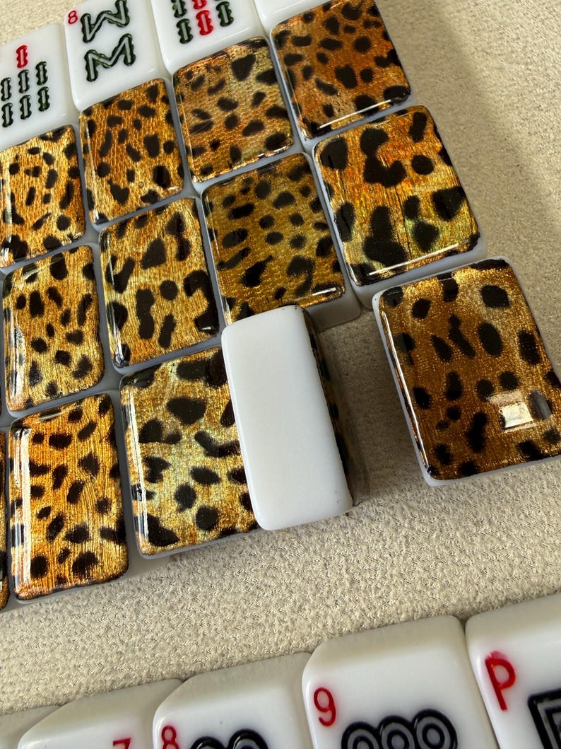 May include: A collection of rectangular mahjong tiles featuring a glossy leopard print pattern in shades of gold, brown, and black. Some tiles are solid white with black and red symbols, including numbers and letters, arranged in a grid formation.