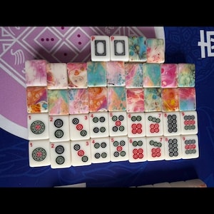 May include: A set of Mahjong tiles arranged on a blue surface. The tiles feature various designs, including colorful abstract patterns and traditional symbols. The tiles are white with black and red markings.