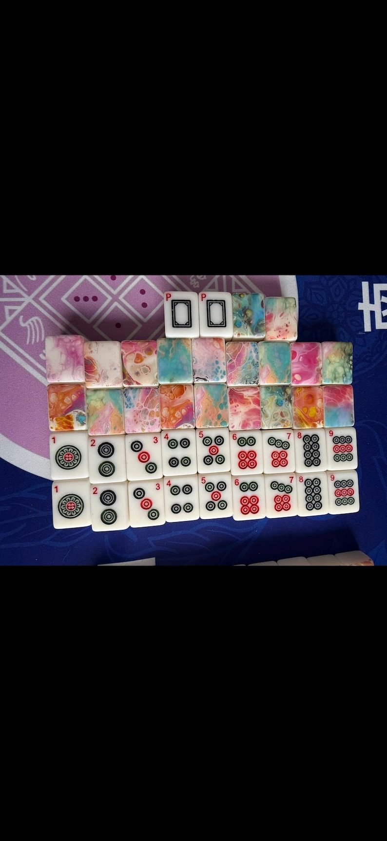 May include: A set of Mahjong tiles arranged on a blue surface. The tiles feature various designs, including colorful abstract patterns and traditional symbols. The tiles are white with black and red markings.