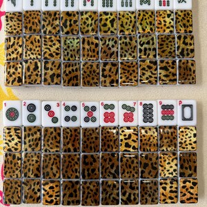 May include: A set of rectangular mahjong tiles featuring a leopard print pattern on the backs. The fronts display various traditional symbols, including bamboo, circles, characters, and the green dragon, with small red numbers 1 through 9 above.