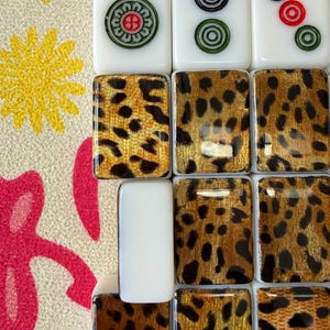 May include: A collection of rectangular mahjong tiles featuring white tops with red numbers 1, 2, and 3, and circular green and black symbols. The backs of the tiles display a glossy gold and black leopard print pattern on a smooth surface.