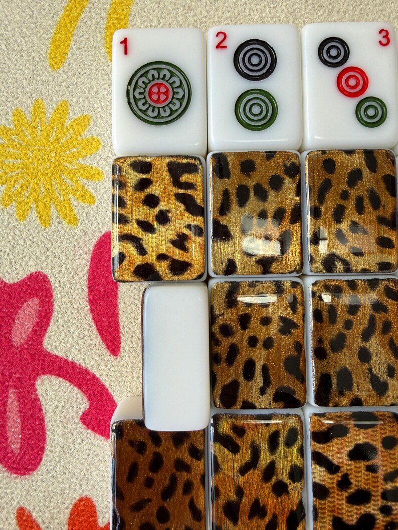 May include: A collection of rectangular mahjong tiles featuring white tops with red numbers 1, 2, and 3, and circular green and black symbols. The backs of the tiles display a glossy gold and black leopard print pattern on a smooth surface.