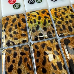 May include: A collection of rectangular mahjong tiles featuring a white base with standard numerical and circular markings on the top row, while the bottom tiles display a glossy, metallic gold and black leopard print pattern on their faces.