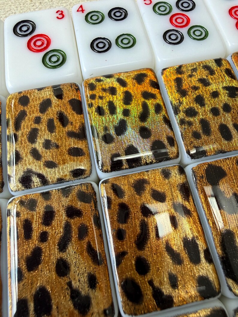 May include: A collection of rectangular mahjong tiles featuring a white base with standard numerical and circular markings on the top row, while the bottom tiles display a glossy, metallic gold and black leopard print pattern on their faces.