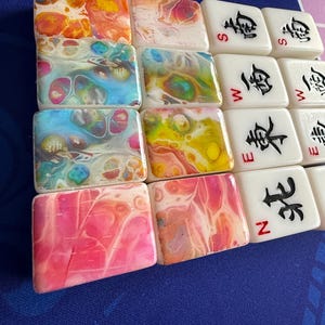 May include: A collection of colorful Mahjong tiles. Some tiles have abstract, swirled patterns in shades of orange, yellow, blue, and pink. Other tiles are white with black Chinese characters and the letters N, S, W, and E.