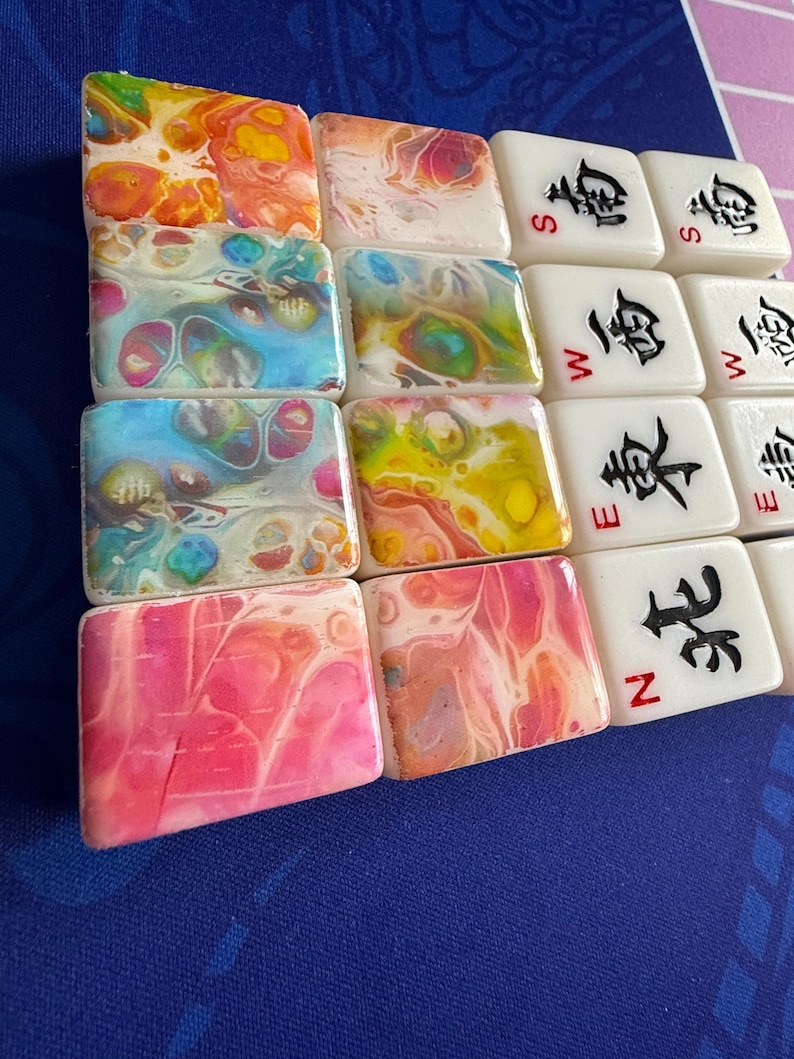 May include: A collection of colorful Mahjong tiles. Some tiles have abstract, swirled patterns in shades of orange, yellow, blue, and pink. Other tiles are white with black Chinese characters and the letters N, S, W, and E.