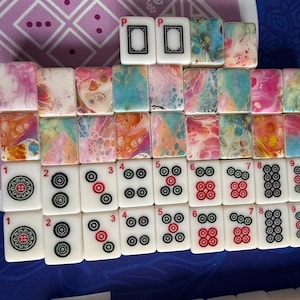 May include: A collection of Mahjong tiles arranged on a purple and blue surface. The tiles feature various designs, including colorful abstract patterns and traditional symbols. The tiles are white with black, red, and green markings.