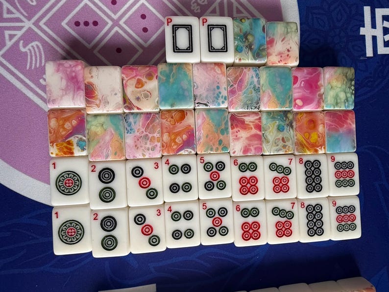May include: A collection of Mahjong tiles arranged on a purple and blue surface. The tiles feature various designs, including colorful abstract patterns and traditional symbols. The tiles are white with black, red, and green markings.