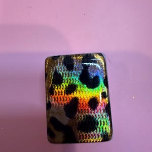 May include: A rectangular cabochon featuring a black leopard print pattern over a vibrant, iridescent, multi-colored background that shifts between shades of gold, orange, green, and blue with a fine, textured, grid-like surface finish.