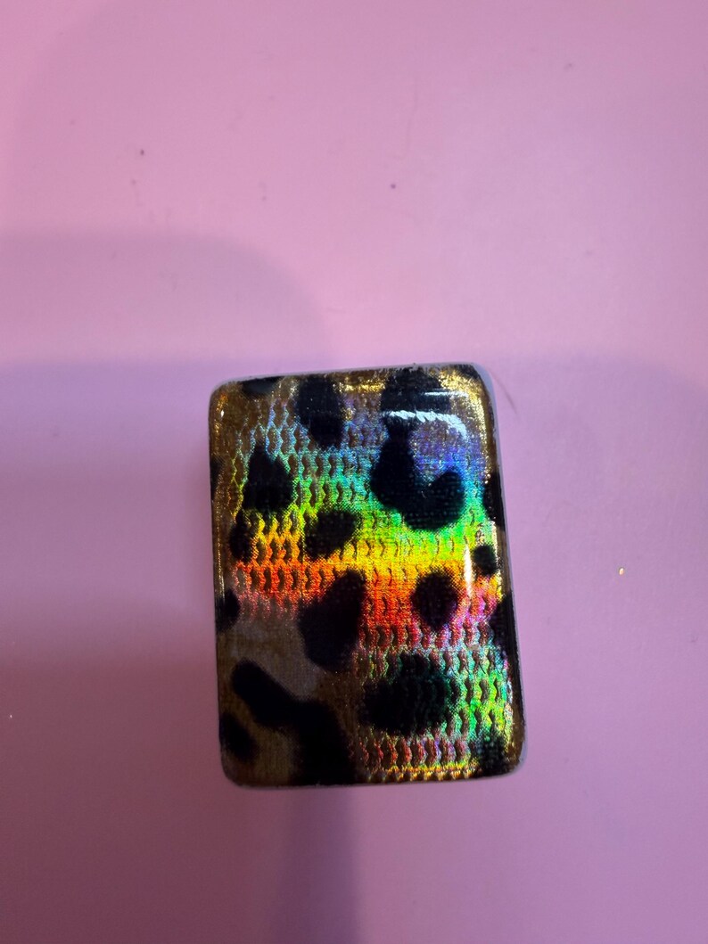 May include: A rectangular cabochon featuring a black leopard print pattern over a vibrant, iridescent, multi-colored background that shifts between shades of gold, orange, green, and blue with a fine, textured, grid-like surface finish.