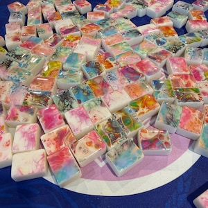May include: A collection of colorful, rectangular soap bars with abstract designs in shades of pink, blue, green, and yellow. The soaps are arranged on a blue surface, creating a vibrant display. The text "DOTS-SOAPS" is visible.