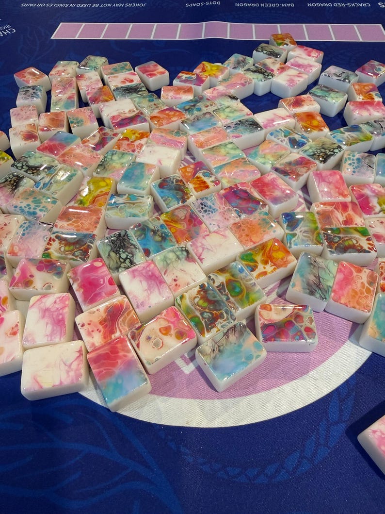 May include: A collection of colorful, rectangular soap bars with abstract designs in shades of pink, blue, green, and yellow. The soaps are arranged on a blue surface, creating a vibrant display. The text "DOTS-SOAPS" is visible.