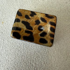 May include: A rectangular brooch featuring a glossy finish with a gold and black leopard print pattern. The surface has a smooth, reflective texture with rounded corners, displaying a repeating animal-inspired spot design across the entire face.