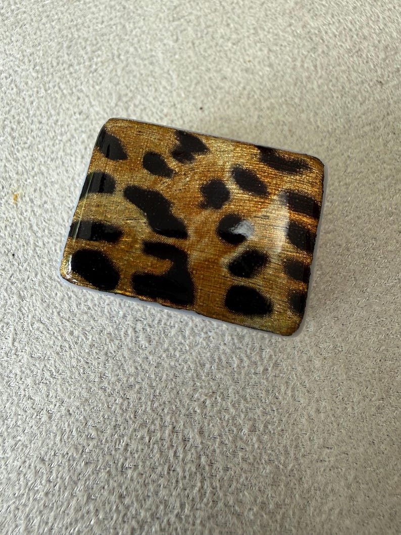 May include: A rectangular brooch featuring a glossy finish with a gold and black leopard print pattern. The surface has a smooth, reflective texture with rounded corners, displaying a repeating animal-inspired spot design across the entire face.