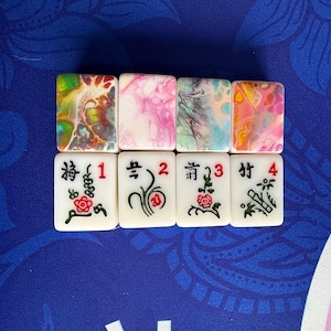 May include: Eight Mahjong tiles. The top row features tiles with colorful abstract designs in shades of yellow, pink, and blue. The bottom row displays white tiles with Chinese characters, numbers, and floral illustrations.