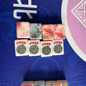 May include: A collection of Mahjong tiles. The top row features tiles with colorful abstract designs in pink, blue, and orange. Below are white tiles with a green design and the word "JOKER" in red.