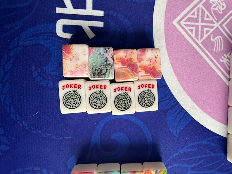 May include: A collection of Mahjong tiles. The top row features tiles with colorful abstract designs in pink, blue, and orange. Below are white tiles with a green design and the word "JOKER" in red.