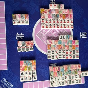 May include: A complete Mahjong set with colorful tiles arranged on a blue playing surface. The tiles feature various symbols and characters, with pink and purple accents. The set includes racks and game instructions.
