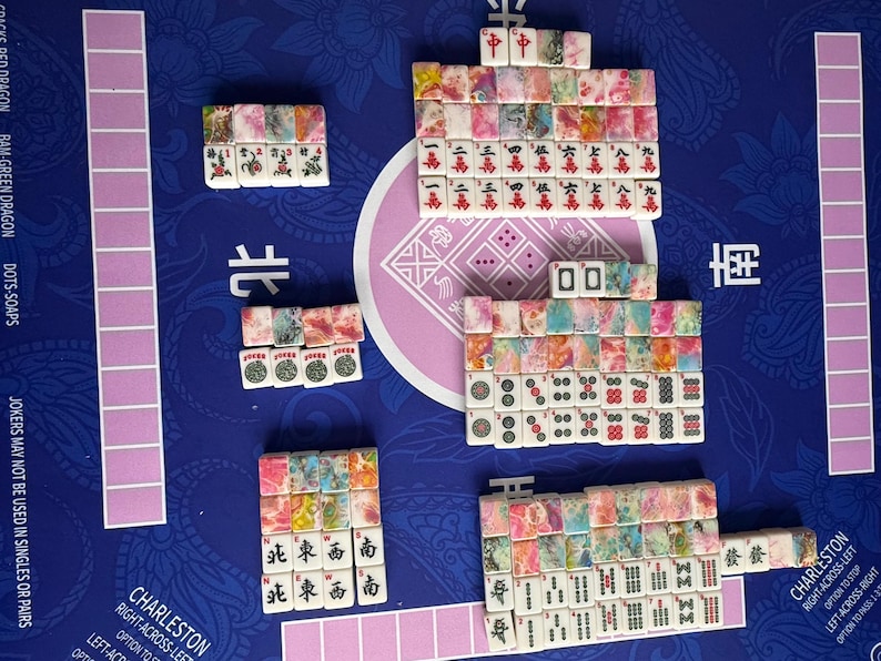 May include: A complete Mahjong set with colorful tiles arranged on a blue playing surface. The tiles feature various symbols and characters, with pink and purple accents. The set includes racks and game instructions.