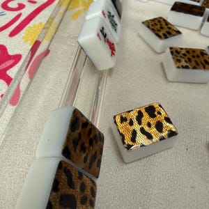 May include: A set of rectangular Mahjong tiles featuring a white base and a gold-toned leopard print pattern on the back. Some tiles are arranged in a clear acrylic rack, while others are scattered, showing red and black character markings.