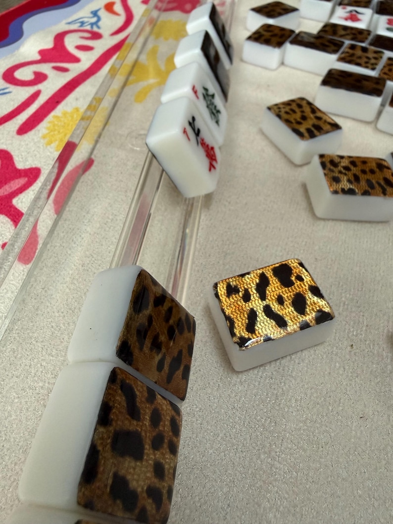 May include: A set of rectangular Mahjong tiles featuring a white base and a gold-toned leopard print pattern on the back. Some tiles are arranged in a clear acrylic rack, while others are scattered, showing red and black character markings.