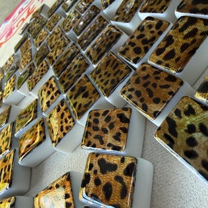 May include: A collection of small, square, white tiles featuring a glossy gold and black leopard print pattern on the top surface. The tiles are arranged in a grid-like formation, showcasing a repeating animal-inspired design on each piece.