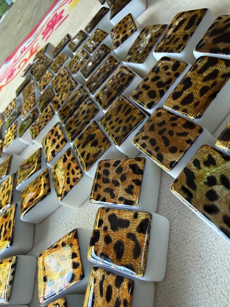 May include: A collection of small, square, white tiles featuring a glossy gold and black leopard print pattern on the top surface. The tiles are arranged in a grid-like formation, showcasing a repeating animal-inspired design on each piece.