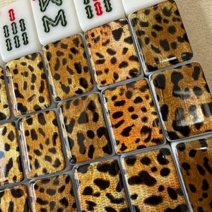 May include: A collection of rectangular mahjong tiles featuring a glossy leopard print pattern in shades of gold, brown, and black. At the top, four white tiles display green and red numerical and character markings, including the number 8 and 9.